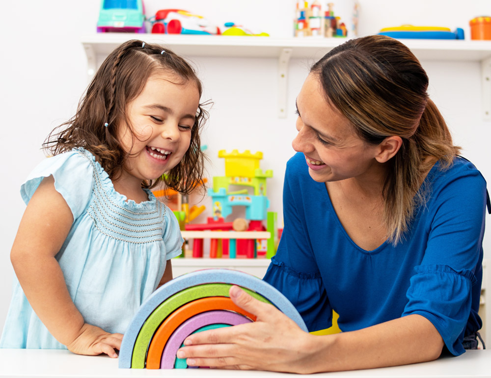 A teacher and a young girl smiling while playing with colorful stacking toys, sharing a joyful moment together.