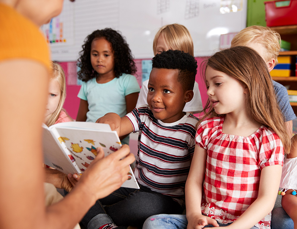 A group of children attentively listening as an teacher reads a book, showing engagement in a classroom environment.