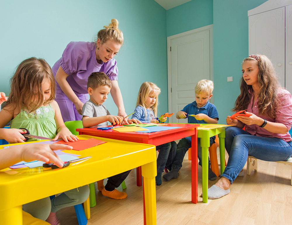 An attentive adult guiding children in a crafting activity, promoting creativity and focus in a vibrant classroom setting.