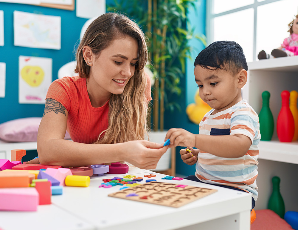 A smiling teacher helps a boy with colorful shapes, creating a joyful learning moment at a bright, playful setting.